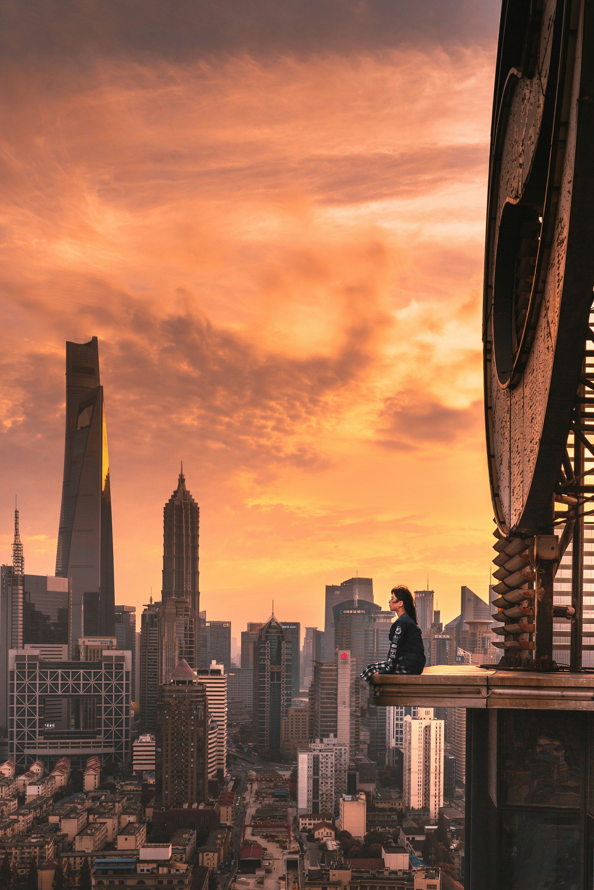 Woman sitting on a rooftop ledge above a city skyline at sunset